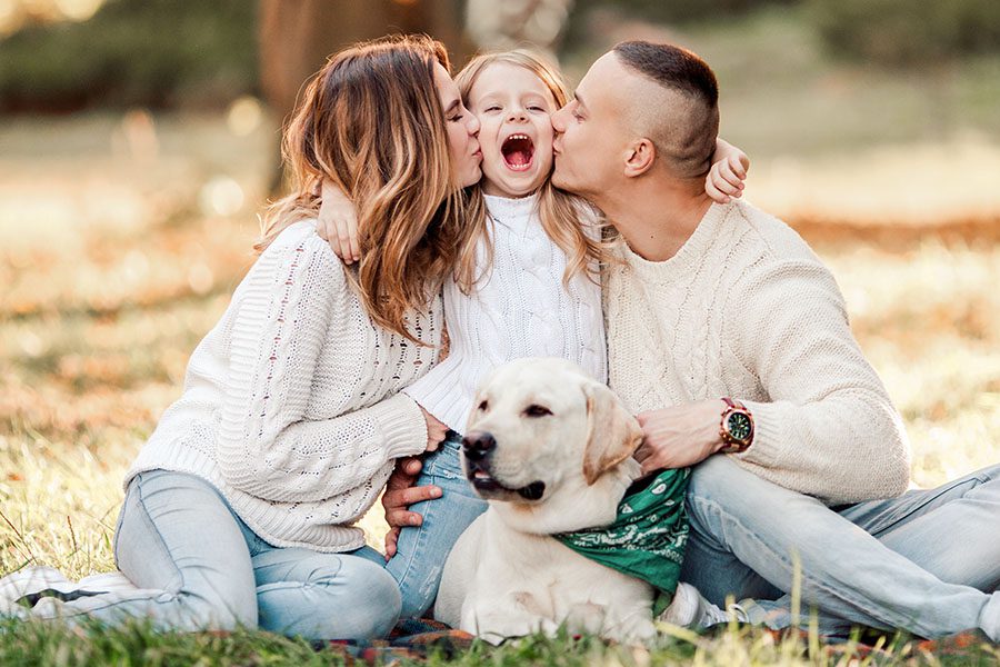 Personal Insurance - Happy Parents Kissing Their Daughter While Sitting Outside On Grass With Dog